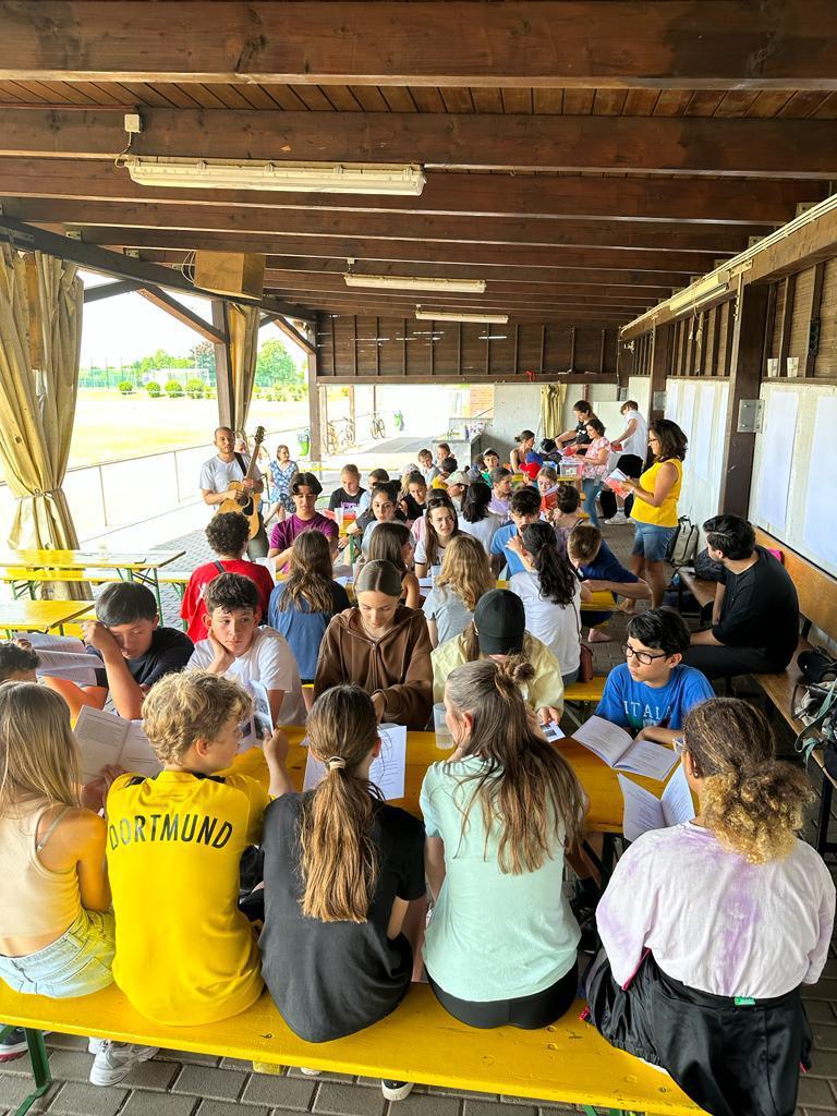 Große Gruppe von Kindern und Jugendlichen an gelben Tischen unter einem Holzpavillon beim Sommercamp, mit Gitarre